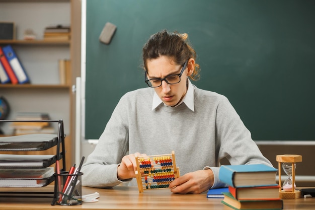 Молодой мыслящий учитель-мужчина в очках держит и смотрит на счеты, сидя за партой со школьными инструментами в классе