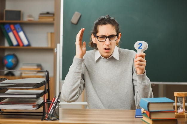 young thinking male teacher wearing glasses sitting at desk holding mathematics numbers with school tools on in classroom