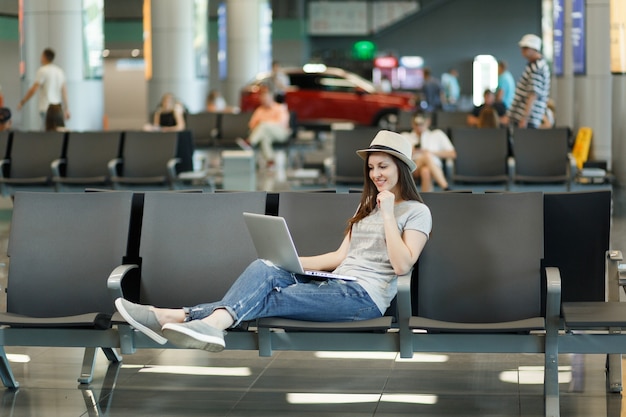 Young thinking traveler tourist woman in hat working on laptop while waiting in lobby hall at international airport