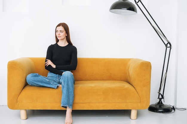 Photo young thinking woman girl with dark long hair in jeans sitting on yellow sofa in the minimalistic interior