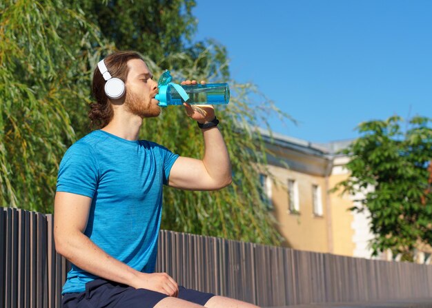 Young thirsty sportsman drinking fresh water from bottle while running outdoors on sunny day