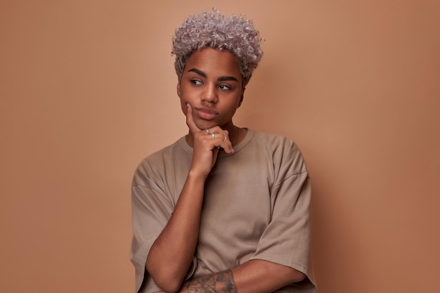 Young thoughtful concentrated African American woman holds hand on chin makes dreamy look and thinks about own future dressed in casual style posing on brown background Planning tomorrow day concept