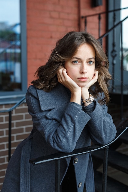 Young thoughtful girl in gray coat and turtleneck poses in front of red building