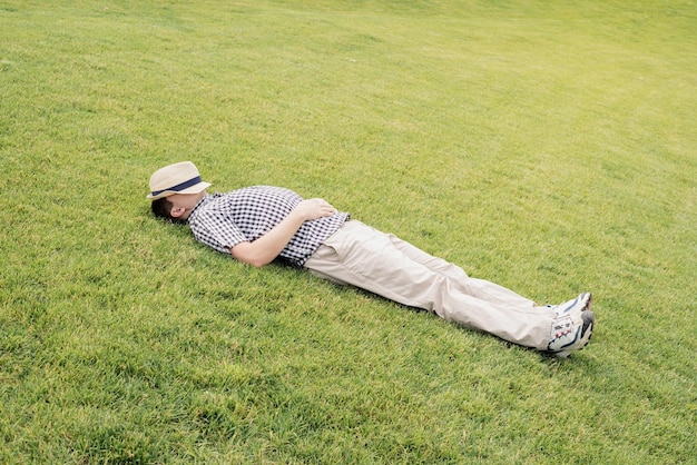 사진 여름 모자를 입은 젊은 사려 깊은 남자가 공원의 잔디에 앉아서 멀리 바라보고 있다.