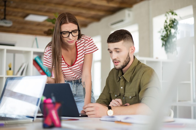 Young thoughtful man in shirt and upset woman in striped Tshirt and eyeglasses working together with laptop Business people spending time at work in modern cozy office