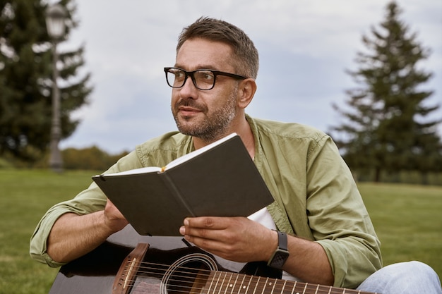 Young thoughtful man wearing eyeglasses holding acoustic guitar\
and composing a song while sitting