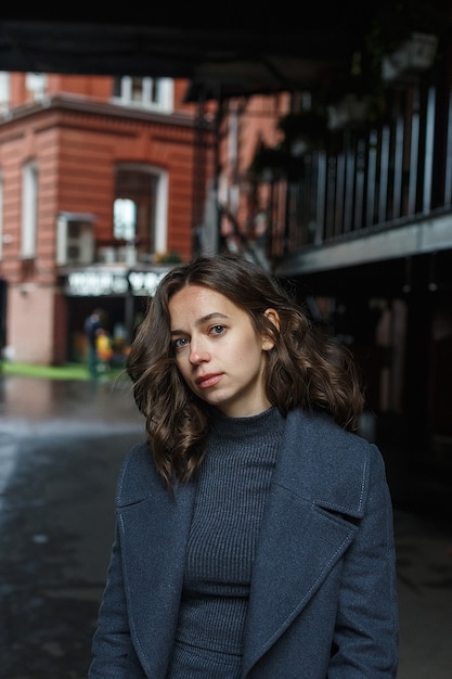 Young thoughtful pretty girl in gray coat and turtleneck poses in front of red building