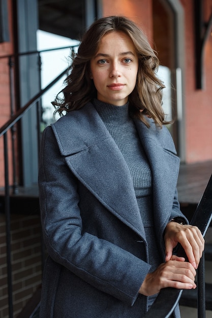 Young thoughtful pretty girl in gray coat and turtleneck poses in front of red building