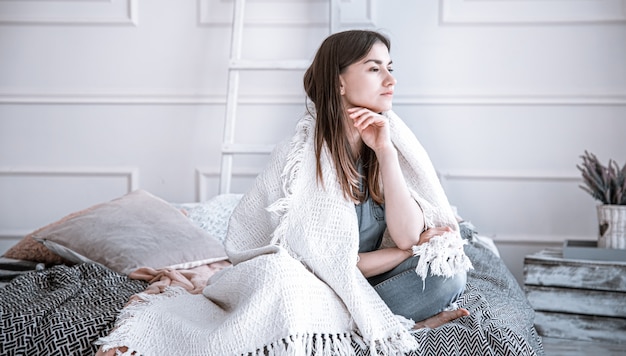 A young, thoughtful woman is sitting on a bed in a cozy room, covered with a blanket in sad mood.