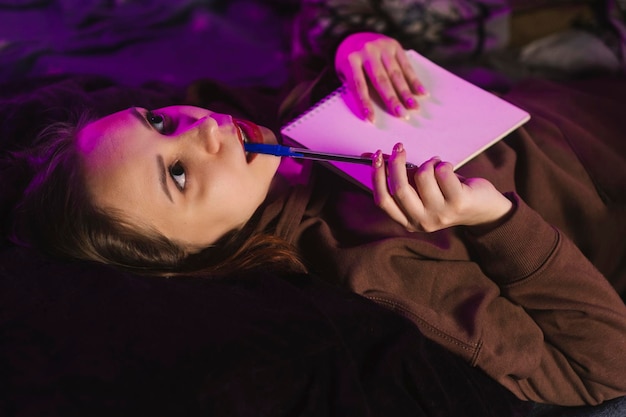 Young thoughtful woman with notepad biting pen lying in bed at night close up relaxed female hugging