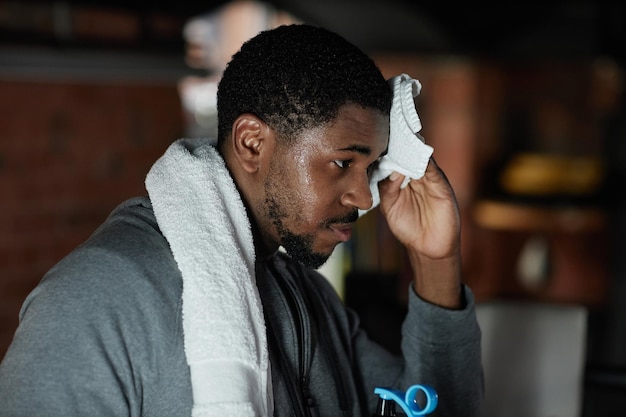 Young tired African American male boxer wiping his wet forehead with white soft towel at break after hard training in gym or sports club