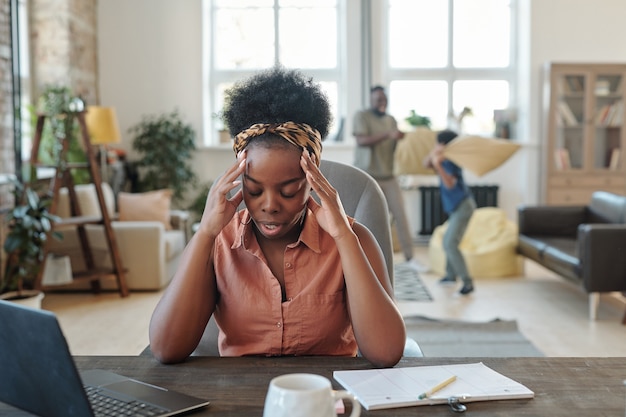 Young tired African woman touching head while sitting by table in front of laptop during network against her husband playing with little son