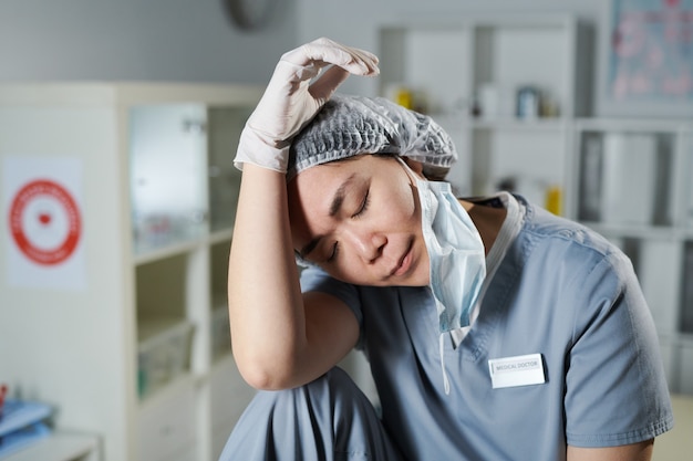 Young tired asian nurse in uniform and protective workwear\
sitting in front of camera and touching her head while keeping eyes\
closed