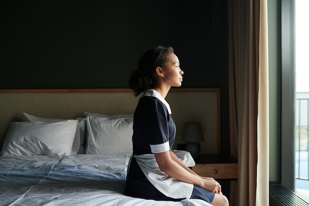 Young tired chambermaid in uniform sitting on double bed in hotel room