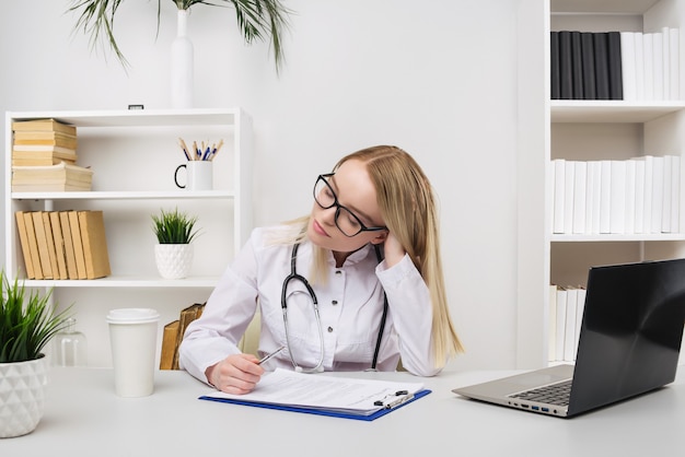 Young tired exhausted woman sitting at desk, working on computer with medical documents in light office in hospital. Female doctor in medical gown sleep in consulting room Healthcare medicine concept