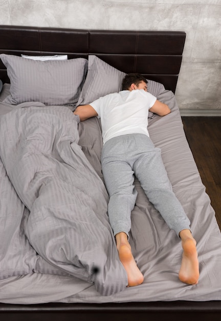 Молодой усталый мужчина в пижаме спит без одеяла в стильной кровати в серых тонах и возле тумбочки со свечами в спальне в стиле лофт