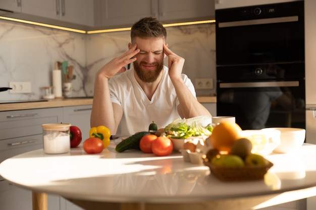 Young tired man with headache sitting at table with vegetables, fruits, eggs and salt. European bearded guy. Interior of kitchen in modern apartment. Concept of healthy nutrition. Sunny daytime