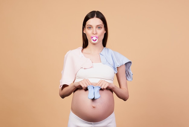 Young tired pretty pregnant girl with a pacifier in her mouth, holding baby blue socks in her hands, baby bodysuits are on her shoulders, preparing for childbirth, preparing for motherhood.