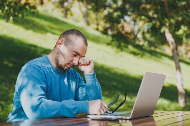 Young tired sad man businessman or student in casual blue shirt, glasses sitting at table with mobile phone in city park using laptop, working outdoors, worries about problems. Mobile Office concept.