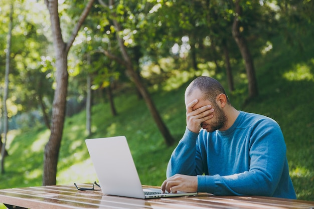 Young tired sad man businessman or student in casual blue shirt, glasses sitting at table with mobile phone in city park using laptop, working outdoors, worries about problems. Mobile Office concept.