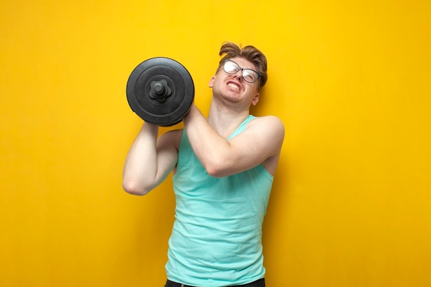 Young tired thin guy nerd lifts heavy dumbbells and cries on yellow background
