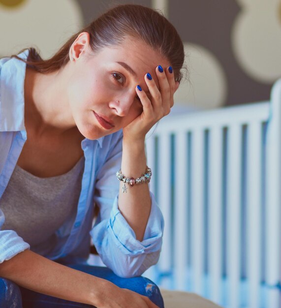 Young tired woman sitting on the bed near children's cot Young mom