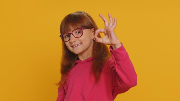 Young toddler school girl in glasses looking approvingly at camera showing okey gesture like sign