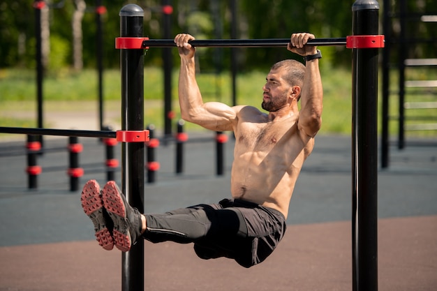 Young topless muscular athlete hanging on sports bar while pulling up on sportsground during workout