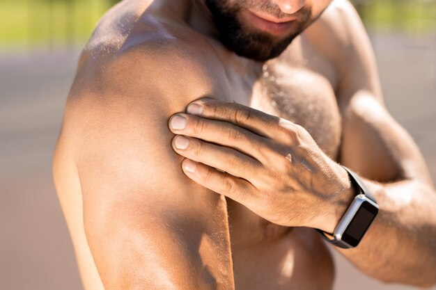 Young topless muscular athlete with smartwatch touching his bicep while training outdoors