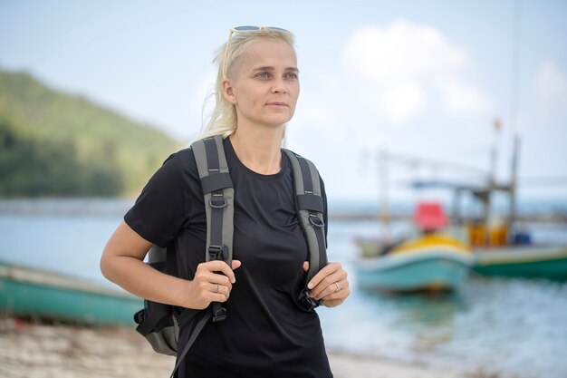 Young tourist of a blonde girl with backpack on the beach with pleasure looking at a beautiful sea landscape, close up