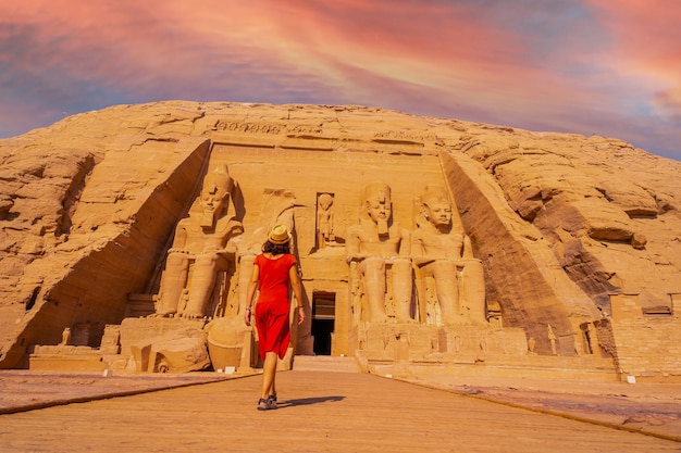 A young tourist girl in red dress walking towards the Abu Simbel Temple in southern Egypt in Nubia next to Lake Nasser at sunset. Temple of Pharaoh Ramses II