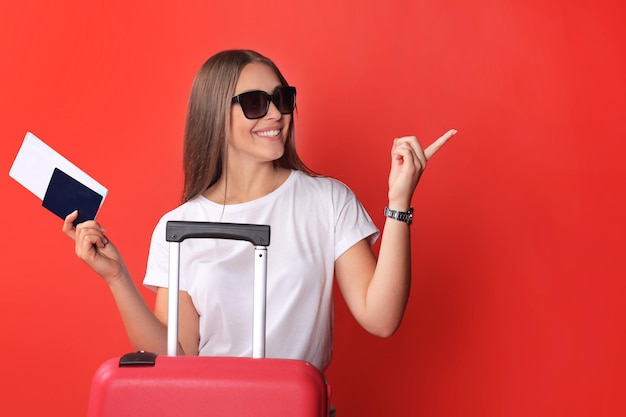 Young tourist girl in summer casual clothes, with sunglasses, red suitcase, passport isolated on red background.