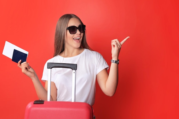 Young tourist girl in summer casual clothes, with sunglasses, red suitcase, passport isolated on red background.