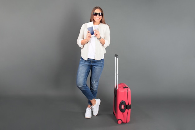 Young tourist girl in summer casual clothes, with sunglasses, red suitcase, passport, tickets isolated grey background.