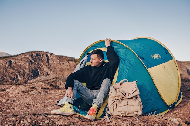 Юный турист поставил в горах палатку и сидит рядом с ней. Рядом лежит походный рюкзак. Отводит взгляд и наслаждается прекрасным видом на холмы.
