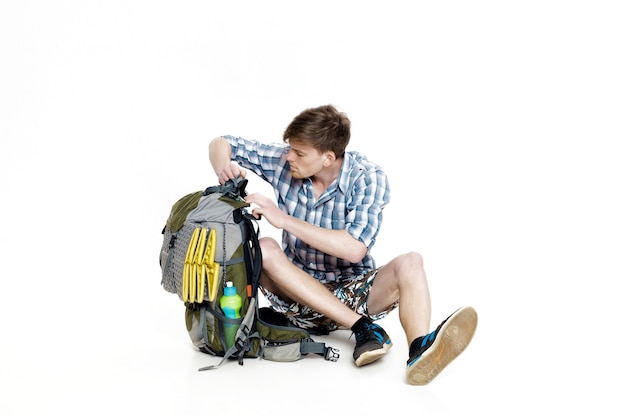 Young tourist is sitting and looking in a backpack something on a white background. The traveler is preparing for a hike