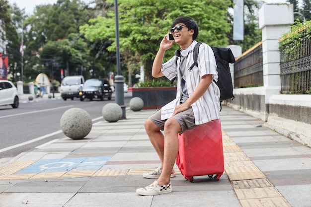 Young tourist man talking on the phone and sitting on luggage at the urban sidewalk