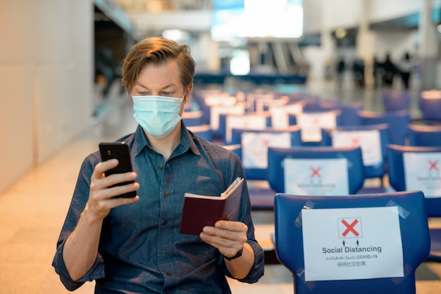 写真 空港で距離を置いて座っている間電話とパスポートをチェックするマスクを持つ若い観光客男