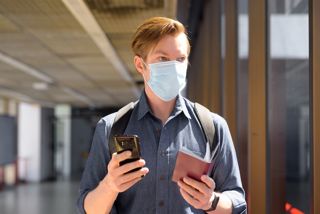 Giovane turista uomo con maschera tenendo il telefono e il passaporto mentre pensa in aeroporto