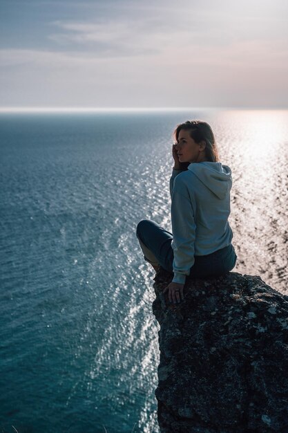 屋外の女性のヨガに座って海の山の風景に沈む夕日を楽しむ若い観光客の女性