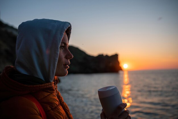 屋外に座って海からの日の出を楽しみながらコーヒータンブラーカップを保持している若い観光客の女性