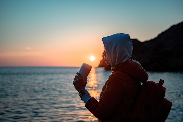 A young tourist woman holding coffee tumbler cup while sitting outdoor and enjoying sunrise over sea