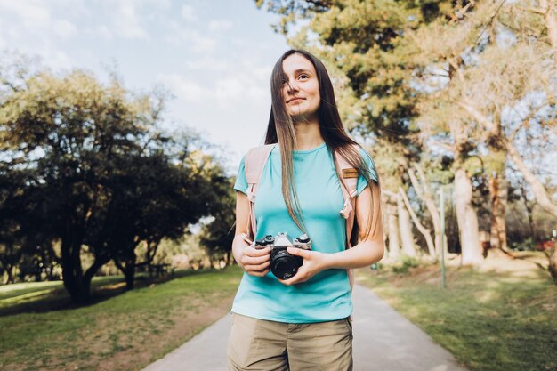 ターコイズのTシャツとバックパックを着て、公園で写真を撮る若い観光客の女性旅行者