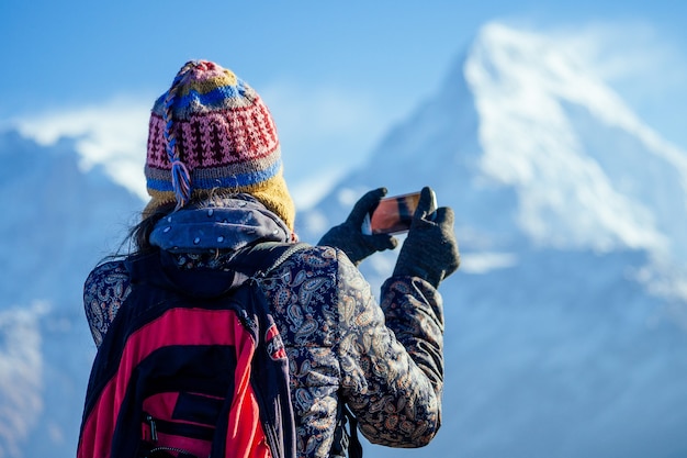 A young tourist woman with a hiking backpack and a knitted hat photographing landscapes in the Himalaya Mountains. trekking concept in the mountains