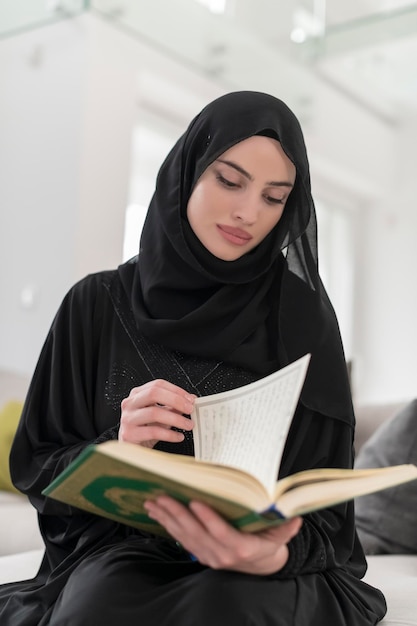 Young traditional muslim woman reading quran on the sofa before iftar dinner during a ramadan feast at home