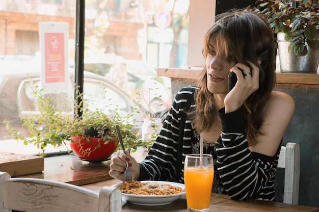 Young transgender woman talking on the phone while eating pasta in a restaurant