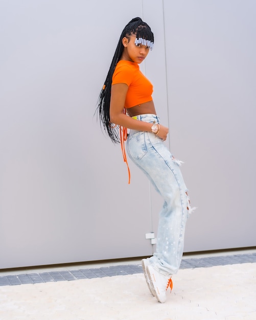 A young trap dancer with braids. Black Breed Girl of African Ethnicity with Orange T-shirt and Cowboy Pants on a Gray Background, Treft PosÃ©