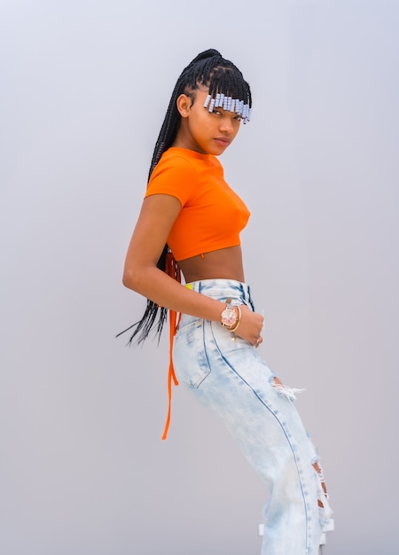 A young trap dancer with braids. Black Breed Girl of African Ethnicity with Orange T-shirt and Cowboy Pants on a Gray Background, Treft PosÃ©