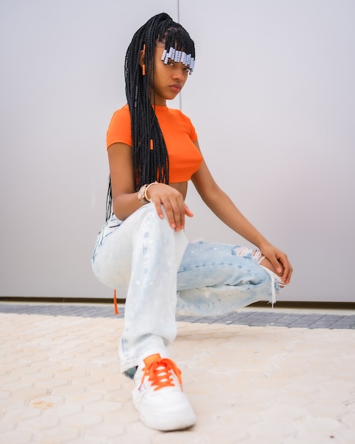 Photo a young trap dancer with braids. black ethnic girl with orange t-shirt and cowboy pants on a gray background, in an urban rapper pose looking at camera