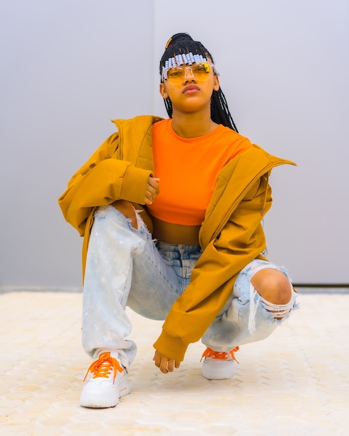 A young trap dancer with braids. Black ethnic girl with t-shirt, orange sunglasses and a jacket, crouched looking at camera with seductive face